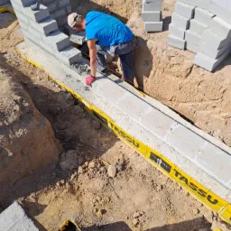 Budowa fundamentów z bloczków betonowych: murarz w niebieskiej koszulce układa zaprawę na pierwszym rzędzie, widoczne wykopane rowy zabezpieczone żółtą folią, stosy bloczków obok.