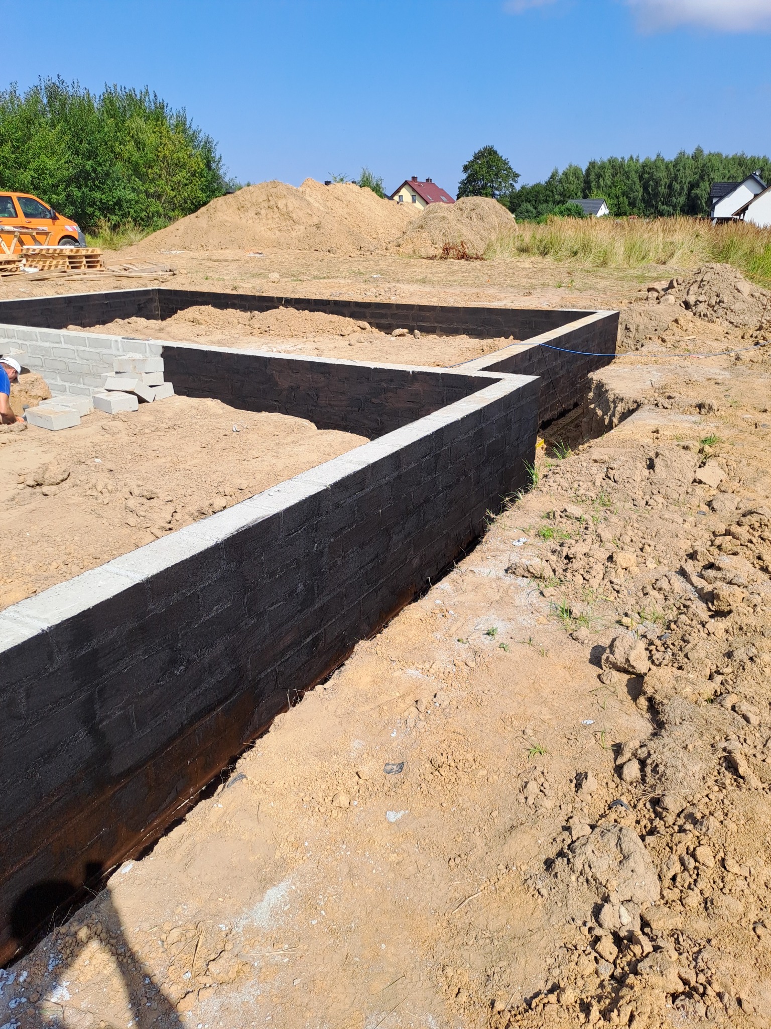 Wykopane fundamenty domu z czarną hydroizolacją, częściowo wymurowane z bloczków betonowych, na działce z hałdą ziemi i widocznym samochodem dostawczym w tle.