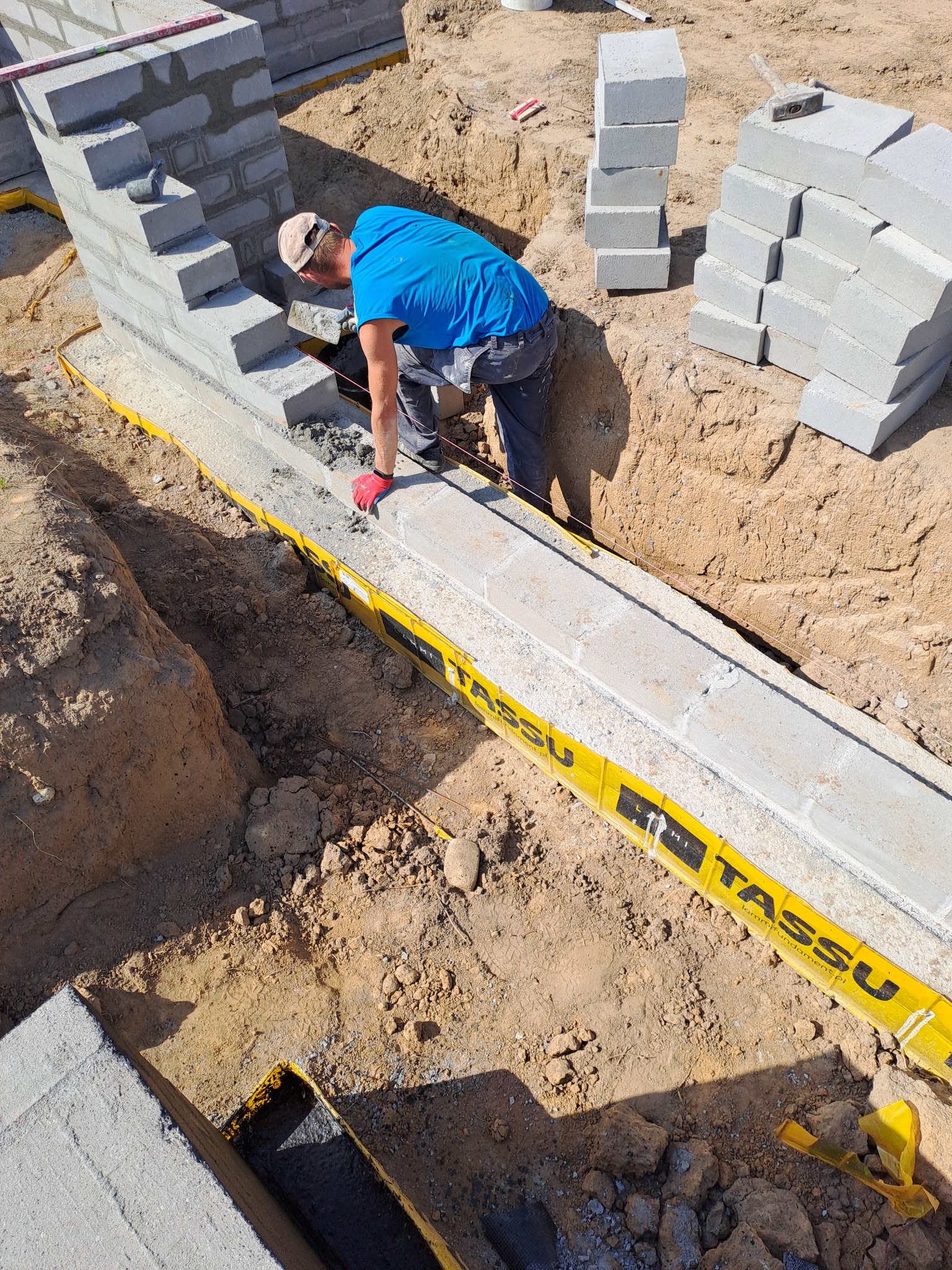 Budowa fundamentów z bloczków betonowych: murarz w niebieskiej koszulce układa zaprawę na pierwszym rzędzie, widoczne wykopane rowy zabezpieczone żółtą folią, stosy bloczków obok.