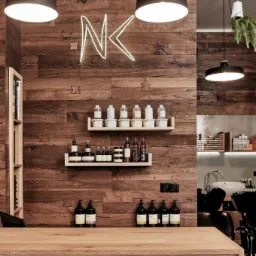 Nowoczesny salon z drewnianą ścianą, neonowym logo NK, półkami z kosmetykami i oświetleniem punktowym, widoczny fragment stanowiska do mycia włosów.