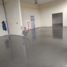 Świeżo wylana, gładka, szara posadzka w surowym wnętrzu z widocznymi instalacjami hydraulicznymi i fragmentami odsłoniętej cegły na ścianie.