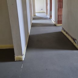 PMF Posadzki Piotr Fesser - Świeżo wylana, szara posadzka w korytarzu budynku w trakcie budowy, z widocznymi dylatacjami i taśmą dylatacyjną przy ścianach. Ściany surowe, częściowo otynkowane, z widoczną cegłą w otworach...