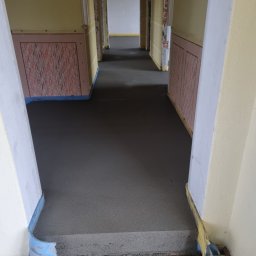 PMF Posadzki Piotr Fesser - Świeżo wylana, ciemnoszara posadzka jastrychowa w długim korytarzu z widocznymi listwami przypodłogowymi zabezpieczonymi niebieską folią.