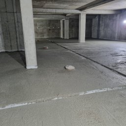 PMF Posadzki Piotr Fesser - Świeżo wylana, mokra posadzka w surowym stanie, z widocznymi listwami poziomującymi i elementami instalacji w podłożu, oświetlona lampami budowlanymi w tle.