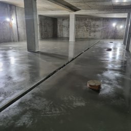 PMF Posadzki Piotr Fesser - Świeżo wylana, mokra posadzka w surowym, betonowym pomieszczeniu z widocznymi dylatacjami i włazami kanalizacyjnymi, oświetlona lampami budowlanymi.