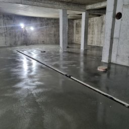 PMF Posadzki Piotr Fesser - Świeżo wylana, błyszcząca posadzka w surowym, betonowym wnętrzu z widocznymi dylatacjami i punktowymi elementami instalacji w podłodze, oświetlona lampami budowlanymi.