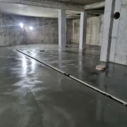 Świeżo wylana, błyszcząca posadzka w surowym, betonowym wnętrzu z widocznymi dylatacjami i punktowymi elementami instalacji w podłodze, oświetlona lampami budowlanymi.