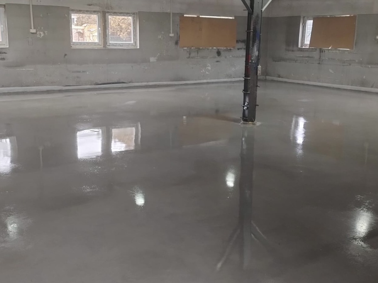 Świeżo wylana, gładka, szara posadzka w surowym wnętrzu z oknami, odbijająca światło, z widocznym słupem konstrukcyjnym w tle.