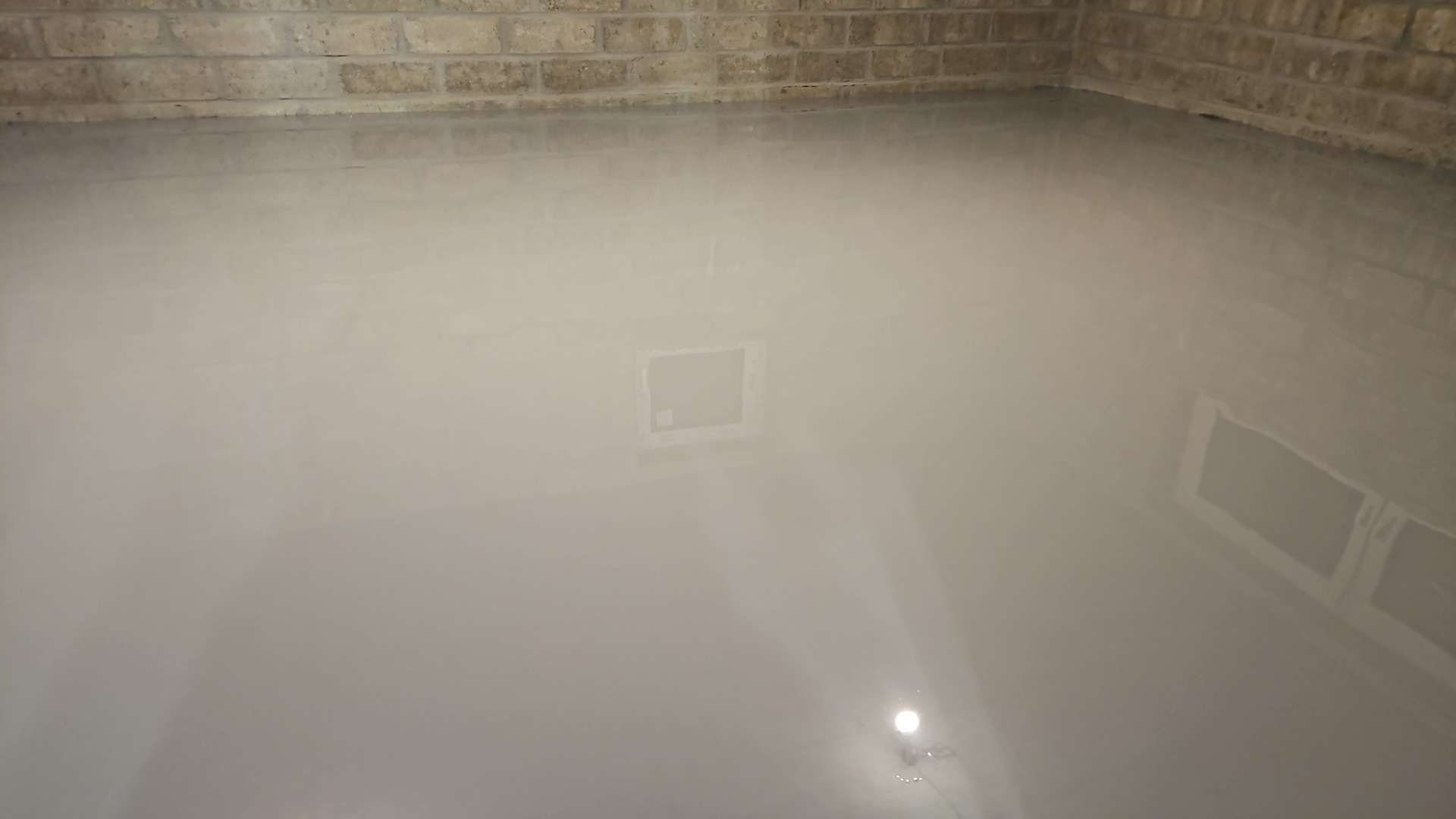 Gładka, szara powierzchnia podłogi z widocznym odbiciem światła, częściowo wykończona ściana z cegły w tle i element wentylacyjny w podłodze.