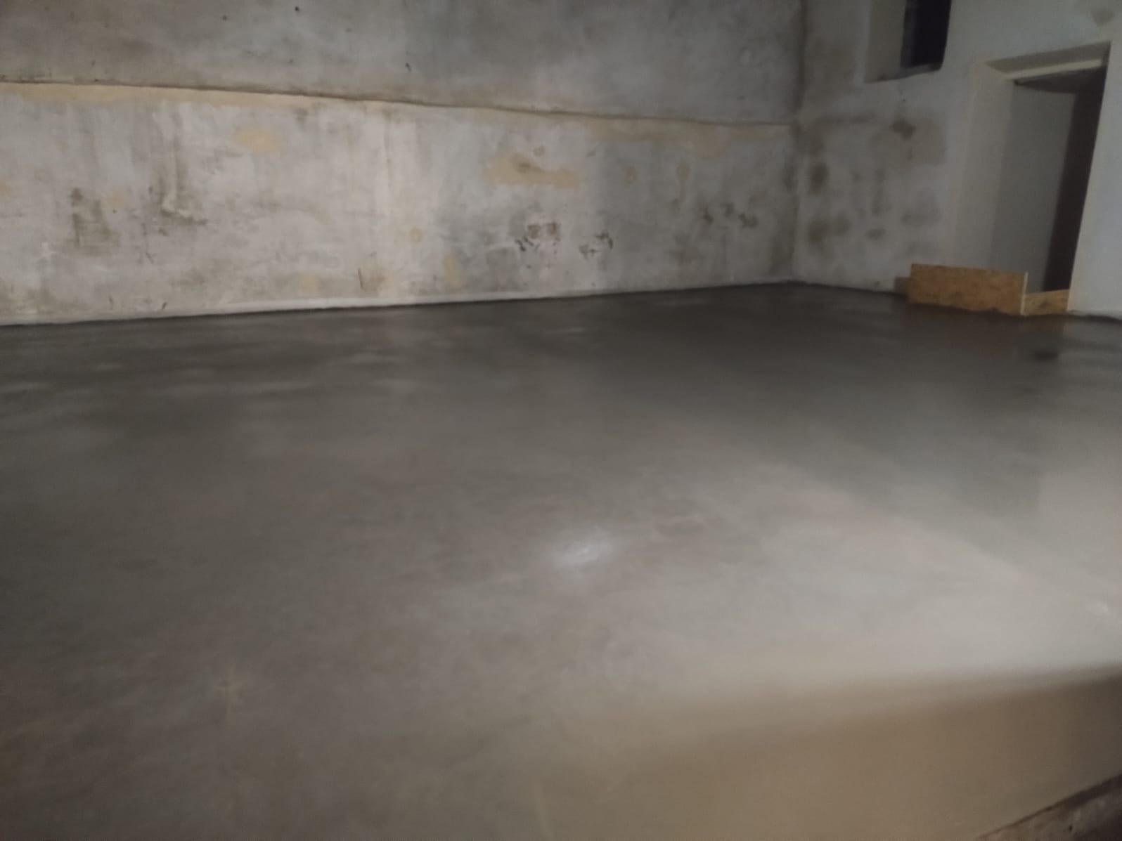 Świeżo wylana, gładka, szara posadzka w surowym pomieszczeniu z betonowymi ścianami. Widoczne ślady po łatach na ścianach.