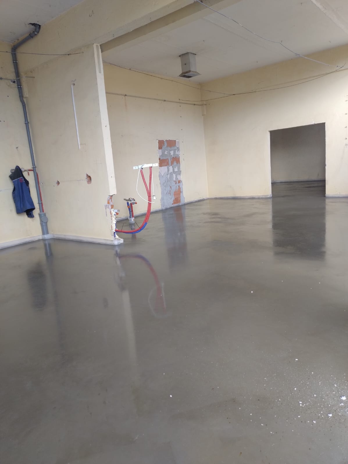 Świeżo wylana, gładka, szara posadzka w surowym wnętrzu z widocznymi instalacjami hydraulicznymi i fragmentami odsłoniętej cegły na ścianie.