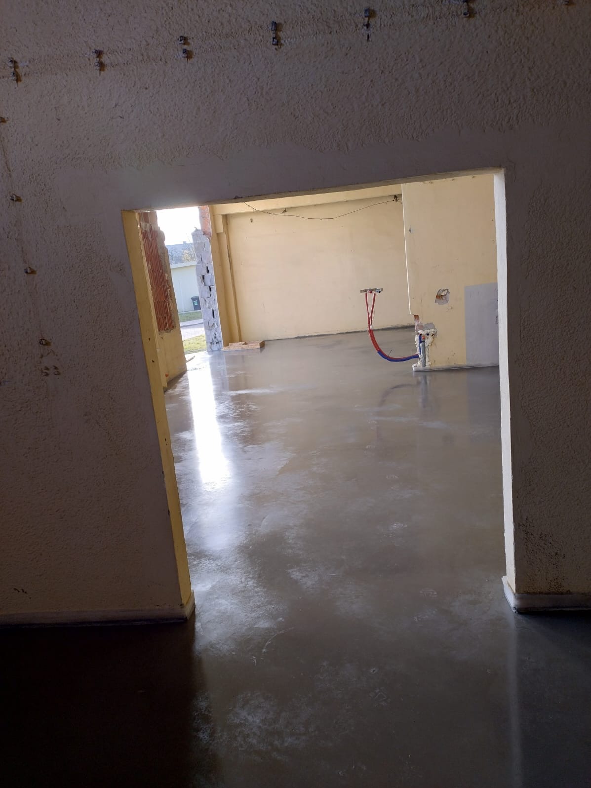 Świeżo wylana, gładka posadzka w surowym stanie, widoczne instalacje hydrauliczne wystające ze ściany, surowy beton na ścianach i suficie.