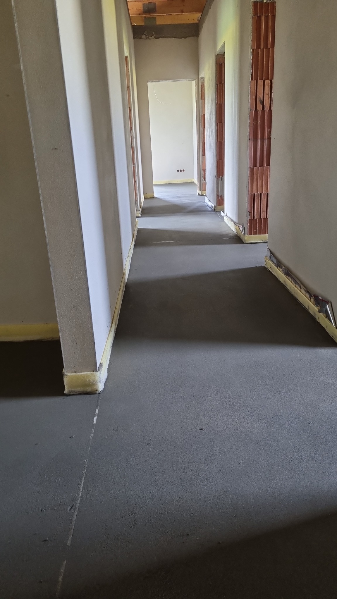 Świeżo wylana, szara posadzka w korytarzu budynku w trakcie budowy, z widocznymi dylatacjami i taśmą dylatacyjną przy ścianach. Ściany surowe, częściowo otynkowane, z widoczną cegłą w otworach...