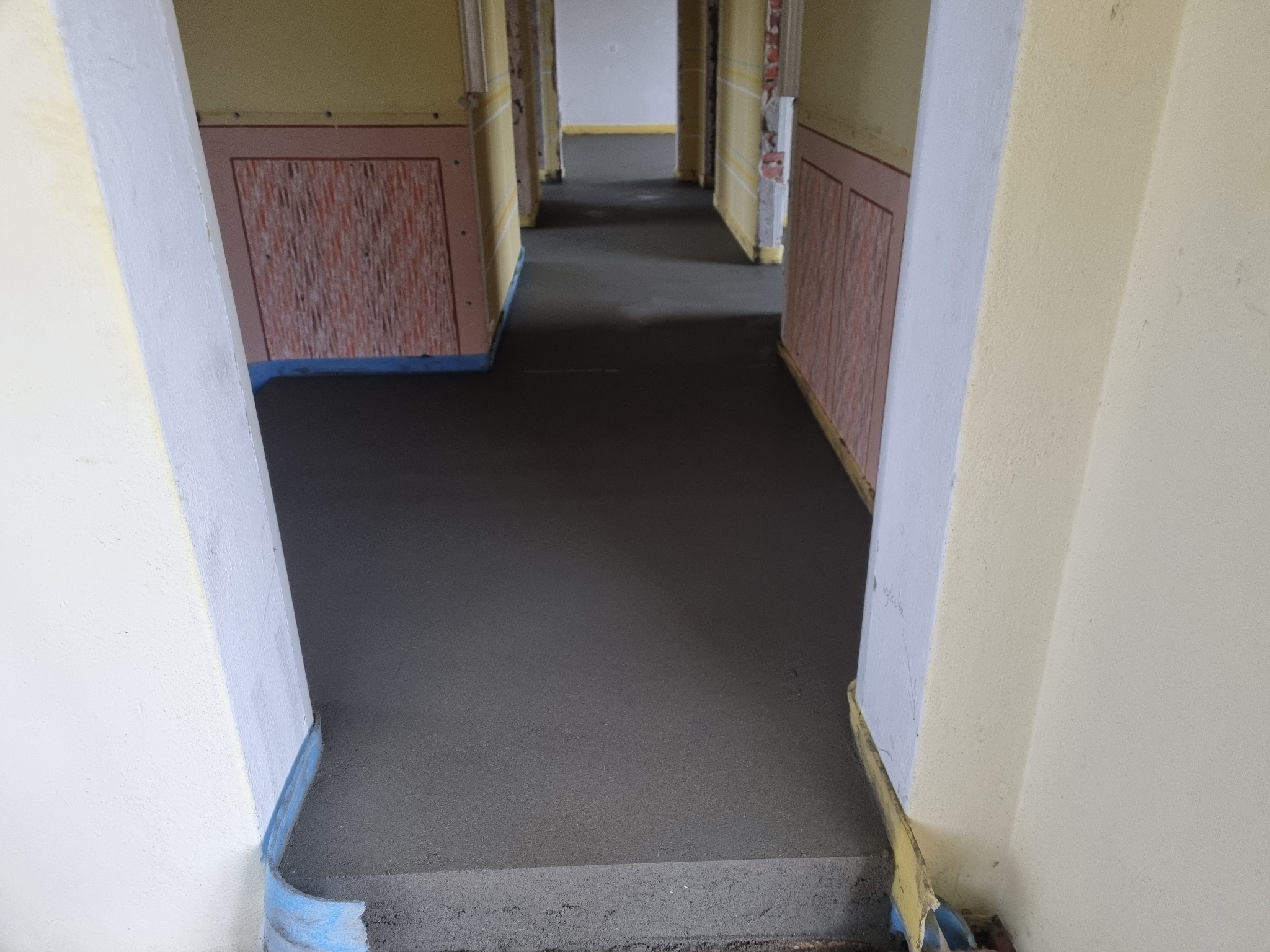 Świeżo wylana, ciemnoszara posadzka jastrychowa w długim korytarzu z widocznymi listwami przypodłogowymi zabezpieczonymi niebieską folią.