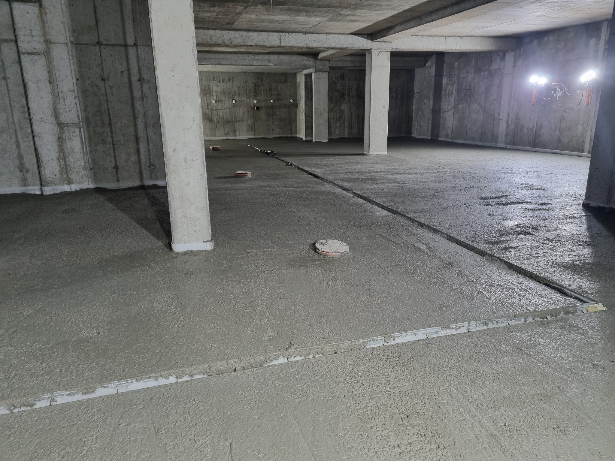 Świeżo wylana, mokra posadzka w surowym stanie, z widocznymi listwami poziomującymi i elementami instalacji w podłożu, oświetlona lampami budowlanymi w tle.
