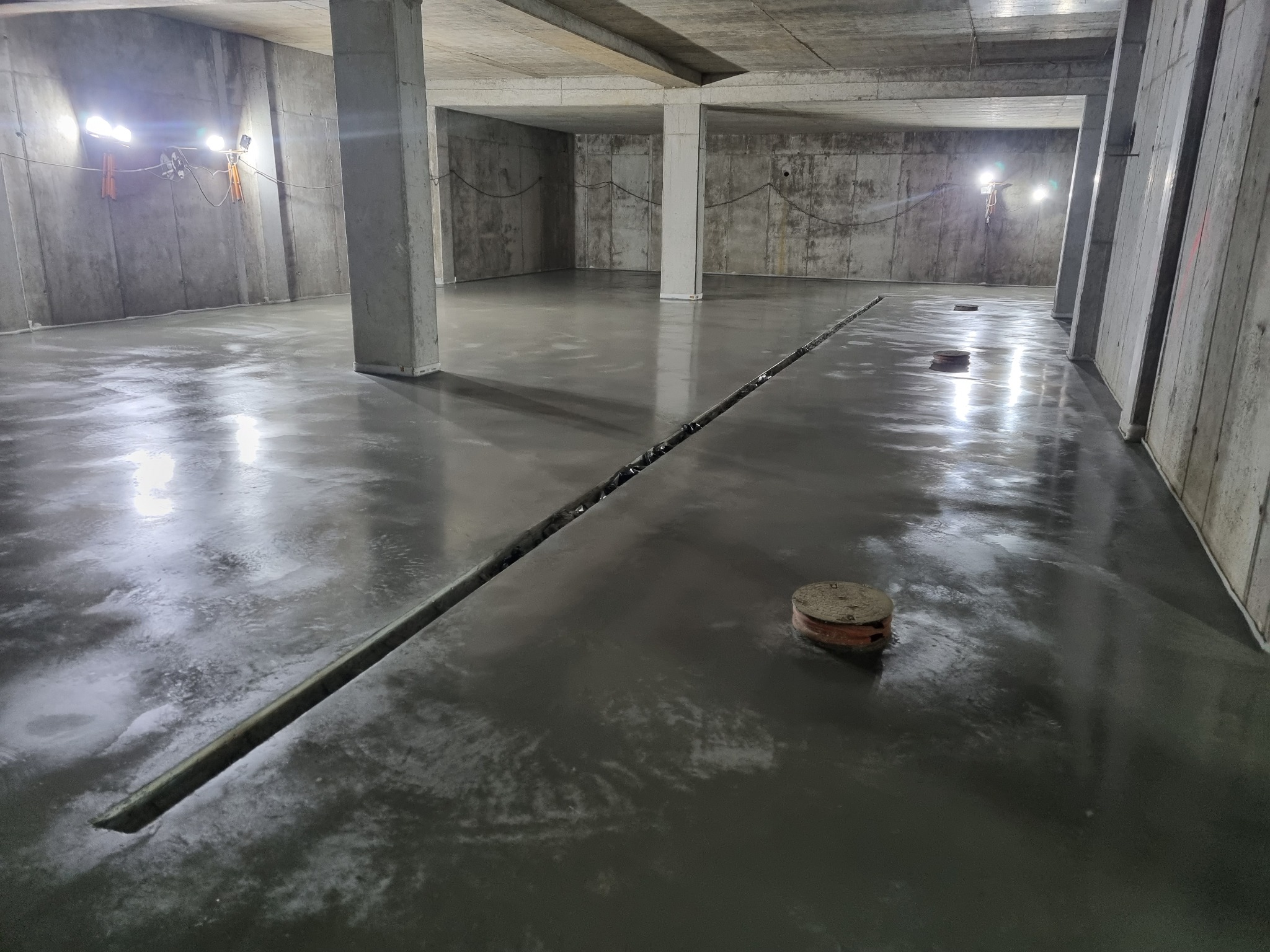 Świeżo wylana, mokra posadzka w surowym, betonowym pomieszczeniu z widocznymi dylatacjami i włazami kanalizacyjnymi, oświetlona lampami budowlanymi.