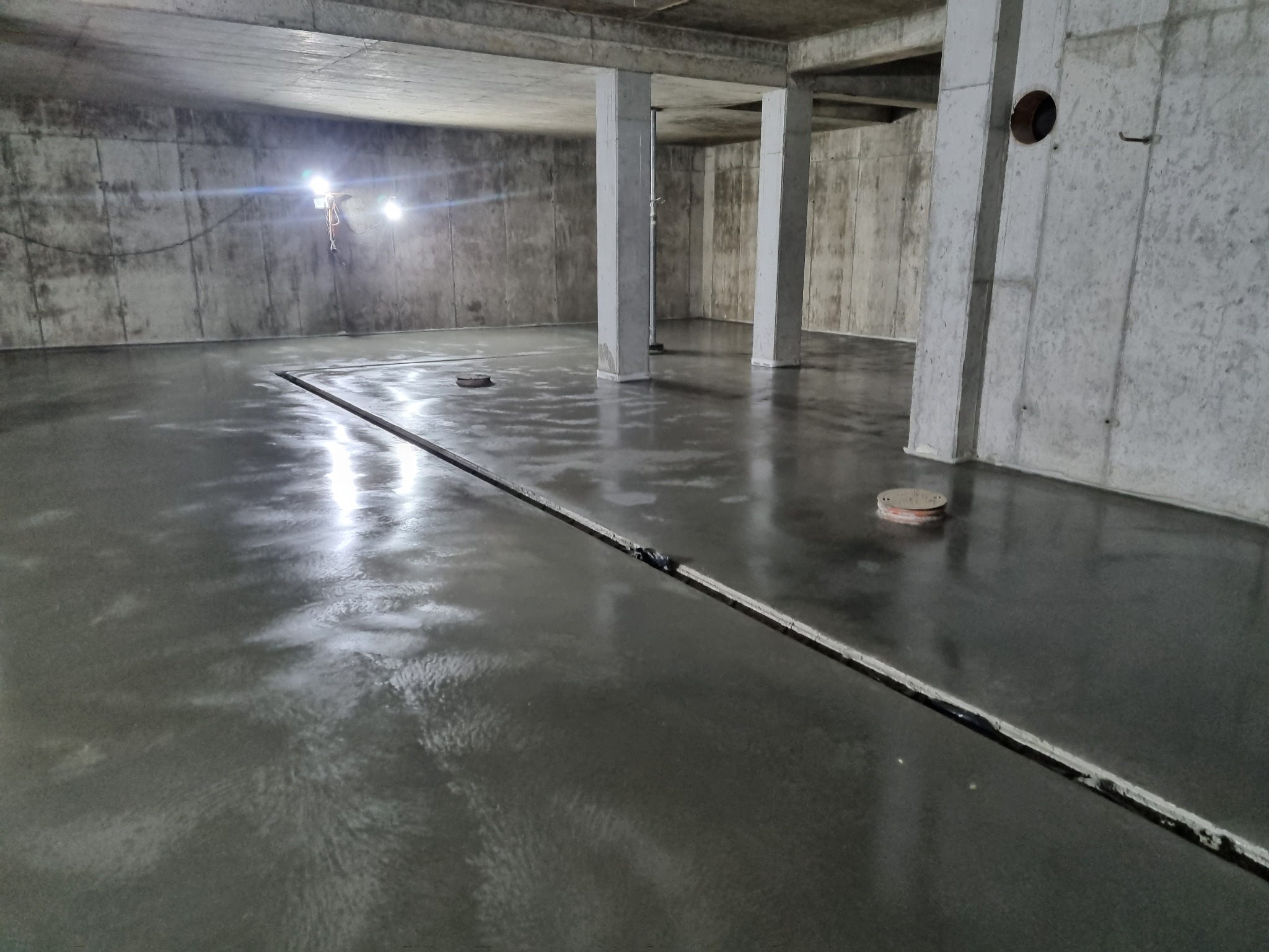 Świeżo wylana, błyszcząca posadzka w surowym, betonowym wnętrzu z widocznymi dylatacjami i punktowymi elementami instalacji w podłodze, oświetlona lampami budowlanymi.