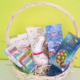 Wiklinowy koszyk wypełniony słodyczami: czekoladki Merci, biały czekoladowy zajączek z czerwoną obrożą, kolorowe ciastka i niebieskie opakowania z czekoladowymi jajkami, na tle jasnozielonej ściany.