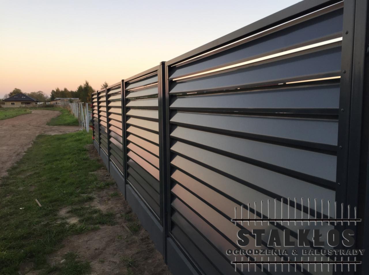 Nowoczesne, antracytowe ogrodzenie z poziomych profili, widoczne z boku, z logo firmy w prawym dolnym rogu, w tle dom i zachodzące słońce.