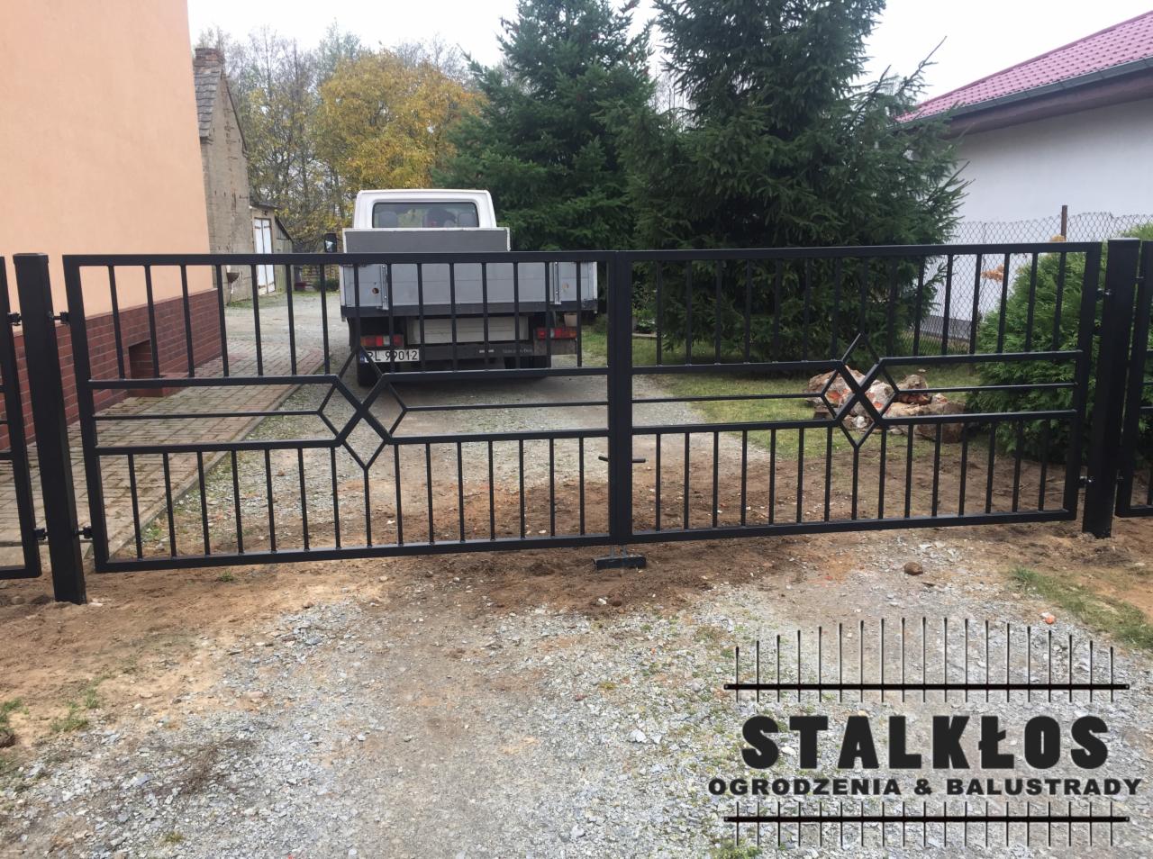 Czarna metalowa brama dwuskrzydłowa z geometrycznymi zdobieniami w kształcie rombów, z widocznym samochodem dostawczym w tle, przed domem z czerwoną cegłą.