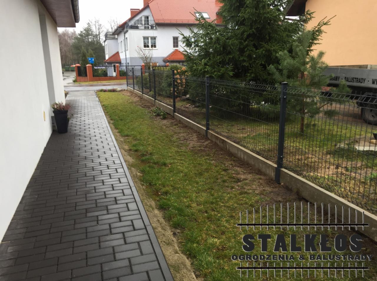 Nowoczesne, ciemnoszare ogrodzenie z siatki panelowej na betonowej podmurówce, oddzielające posesję z kostką brukową od trawnika, w tle dom z czerwoną dachówką i duża choinka.