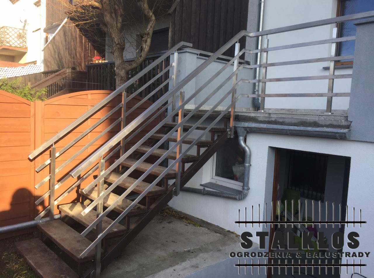 Zewnętrzne schody z drewnianymi stopniami i metalową balustradą, prowadzące do drzwi wejściowych budynku. Widoczny brązowy płot w tle i logo firmy w prawym dolnym rogu.