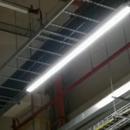 Metalowe korytka kablowe pod sufitem z widocznymi przewodami elektrycznymi w kolorach żółtym, czerwonym i czarnym, oświetlone lampą jarzeniową.