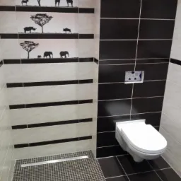 Nowoczesna łazienka z czarno-białymi płytkami i motywem afrykańskich zwierząt na ścianie, widoczny odpływ liniowy pod prysznicem i podwieszana toaleta.