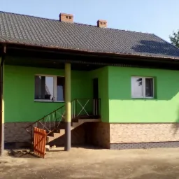 Dom jednorodzinny z jasną, zieloną elewacją, kamienną podmurówką i ciemnym dachem z dwoma kominami, widoczny z zewnątrz w słoneczny dzień.