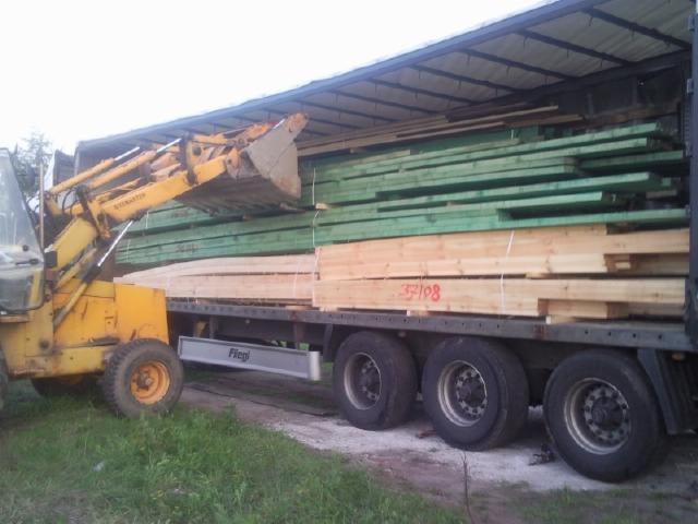 Żółta ładowarka załadowuje drewno budowlane, część pomalowana na zielono, na naczepę ciężarówki. Widoczny numer 37108 na boku naczepy.