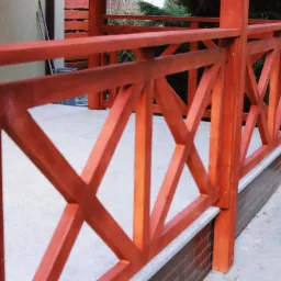 Drewniana balustrada tarasu w kolorze terakoty z krzyżowym wzorem, wsparta na murku z cegły klinkierowej, z widocznym systemem rynnowym.