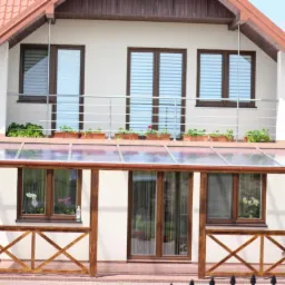 Zadaszony taras przy domu z balustradą balkonową, widoczne rośliny w donicach i drewniane elementy konstrukcyjne.