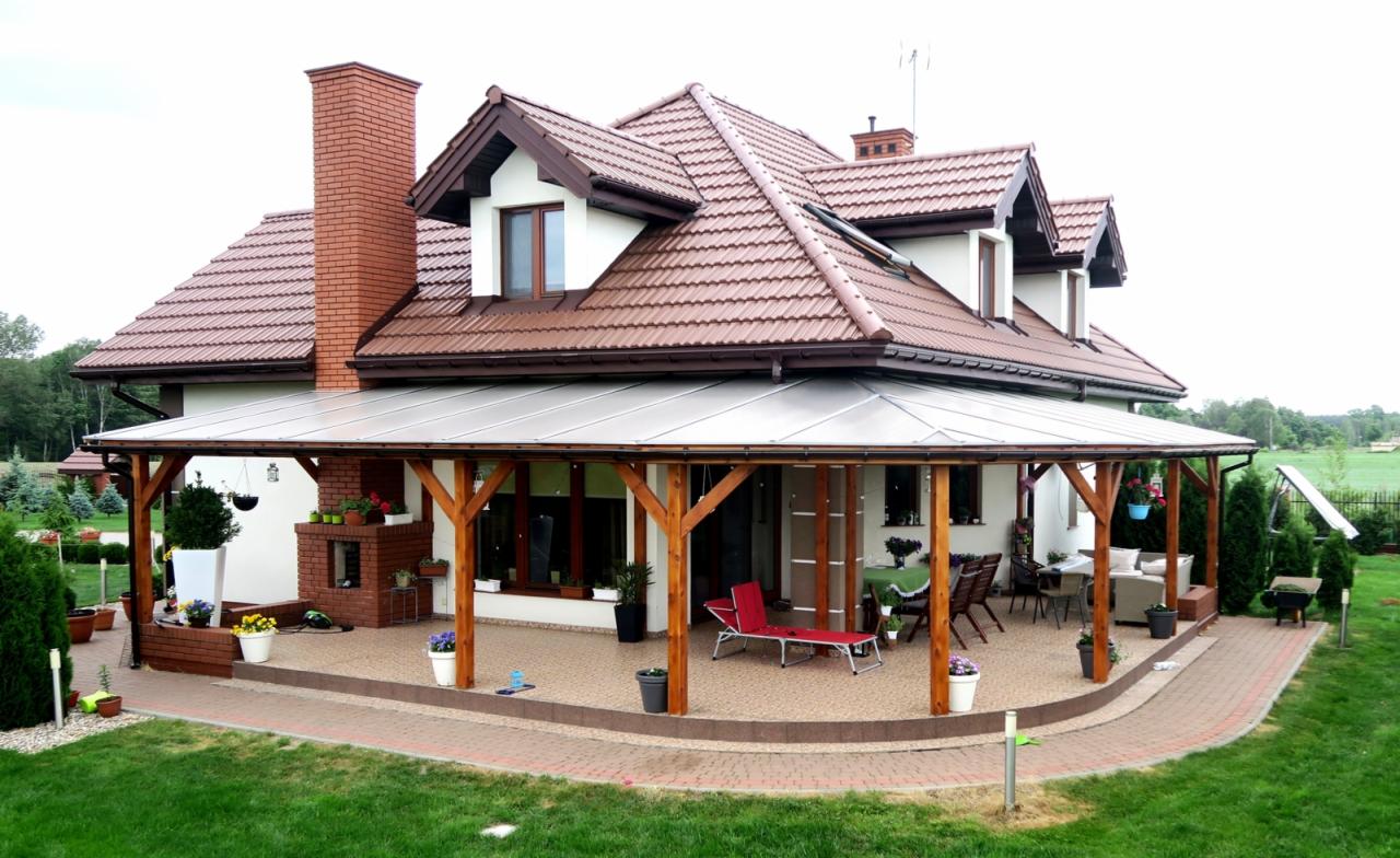 Zadaszony taras przylegający do domu z ceglanym kominem, drewnianą konstrukcją wsporczą i poliwęglanowym pokryciem, otoczony zadbanym ogrodem z trawnikiem i ścieżką z kostki.