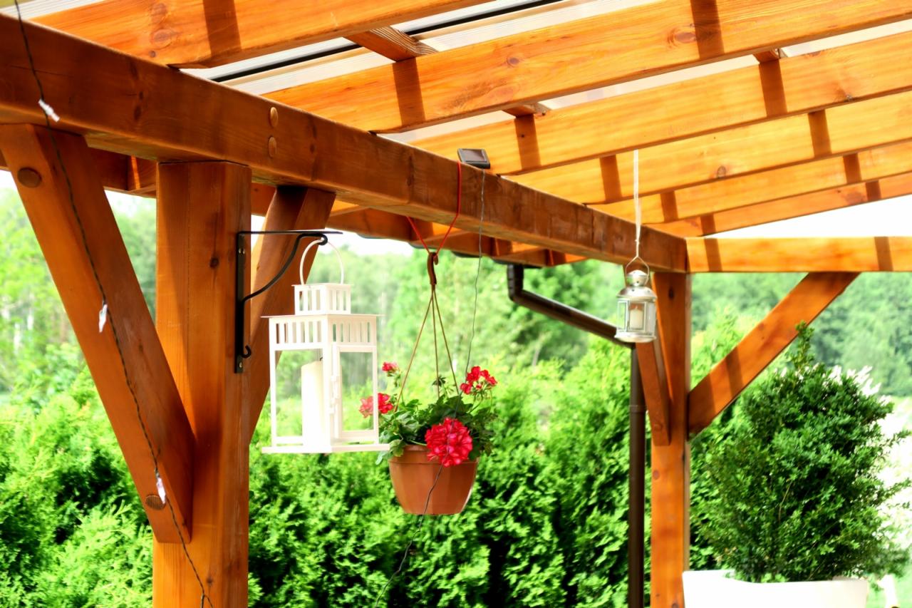 Drewniana konstrukcja z zadaszeniem z poliwęglanu, ozdobiona białymi lampionami i wiszącymi kwiatami, otoczona zielenią.