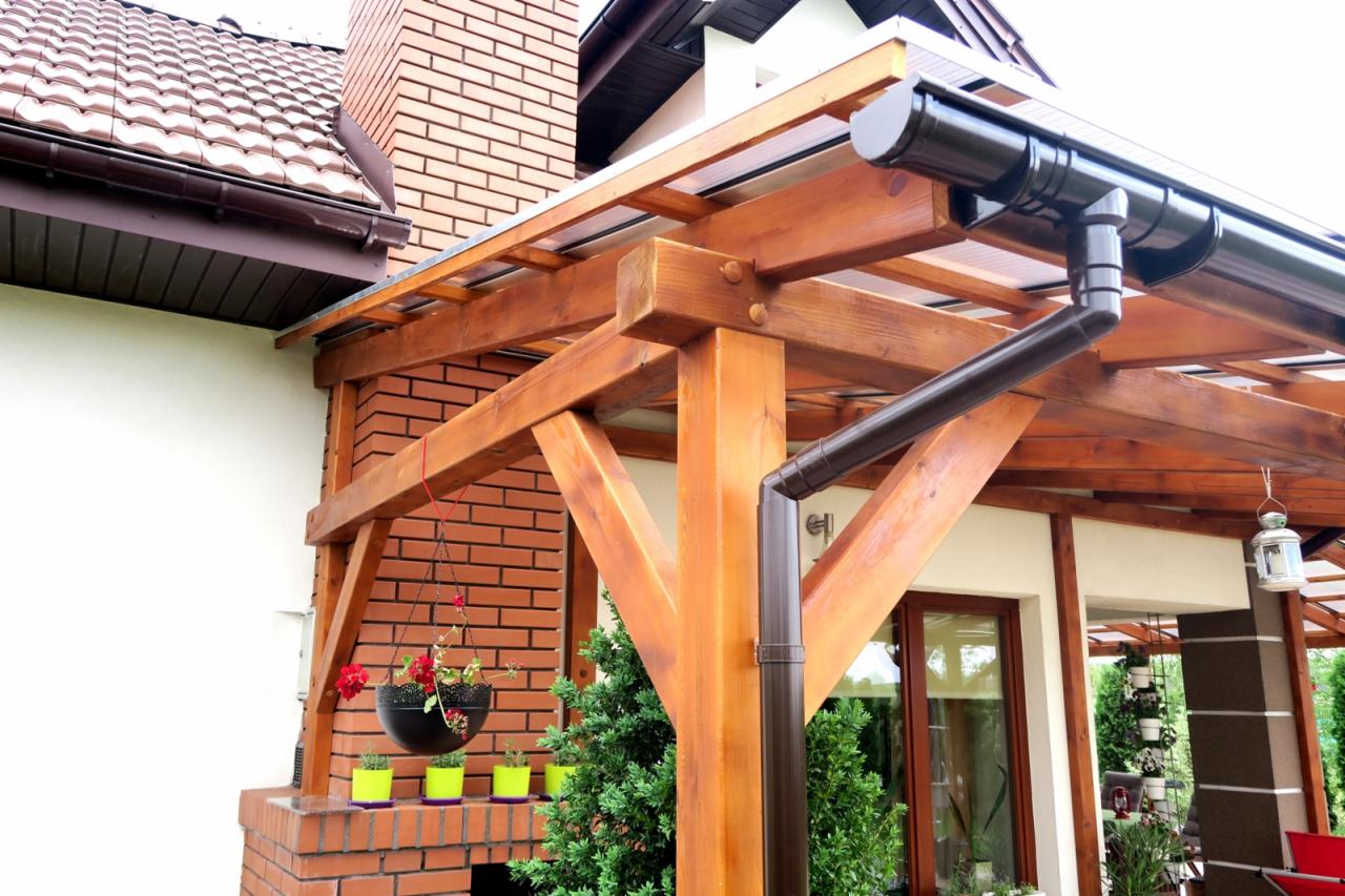 Drewniana konstrukcja z zadaszeniem z poliwęglanu przylegająca do domu, z widocznym systemem rynnowym, wiszącą doniczką i rzędem małych doniczek na ceglanej podstawie.