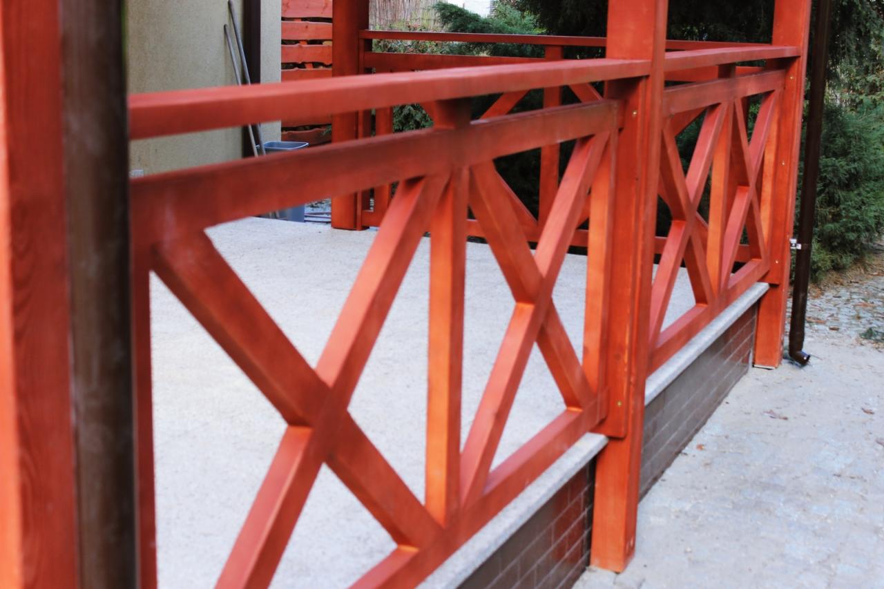 Drewniana balustrada tarasu w kolorze terakoty z krzyżowym wzorem, wsparta na murku z cegły klinkierowej, z widocznym systemem rynnowym.