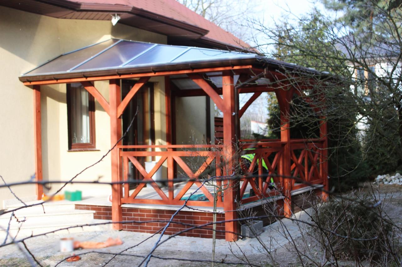 Zadaszony taras z drewnianą konstrukcją i poliwęglanowym dachem, widoczny z zewnątrz, z elementami dekoracyjnymi w postaci krzyżujących się desek na balustradzie.