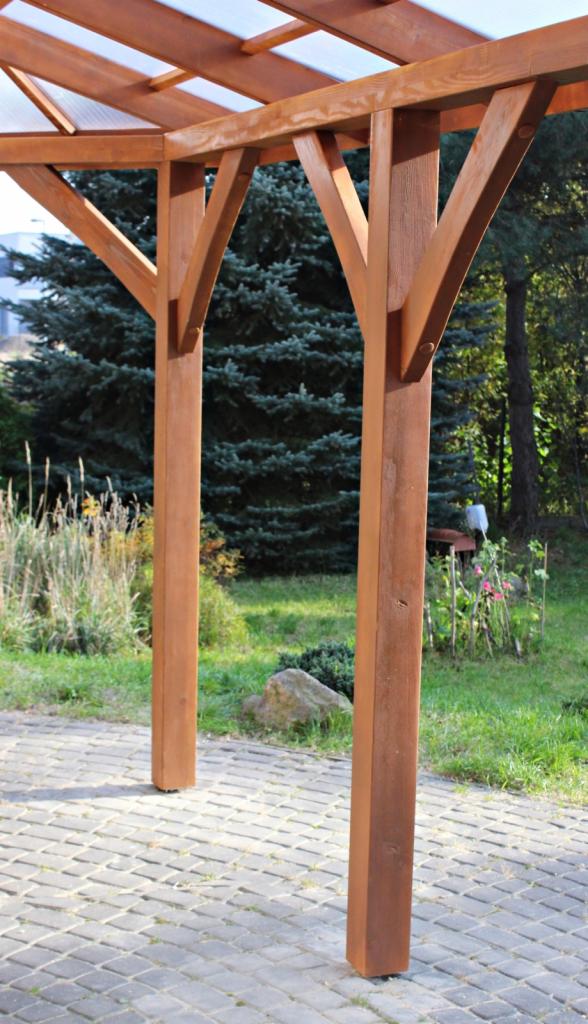 Drewniana konstrukcja z zadaszeniem z poliwęglanu, widok z dołu na tle zieleni ogrodu i brukowanej ścieżki.