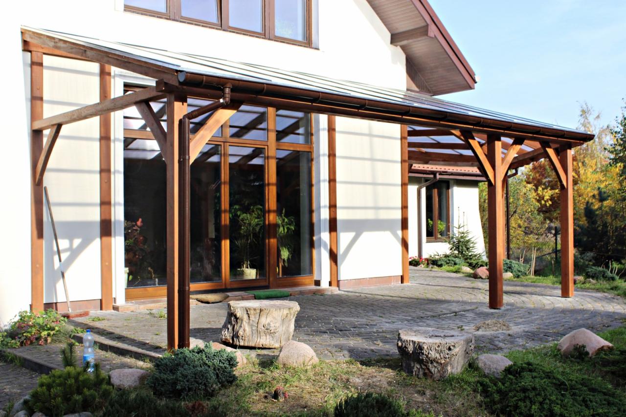 Zadaszony taras z drewnianą konstrukcją i przezroczystym poliwęglanowym dachem, widok z zewnątrz na tle domu z dużymi oknami i zadbanym ogrodem z kamienną ścieżką.