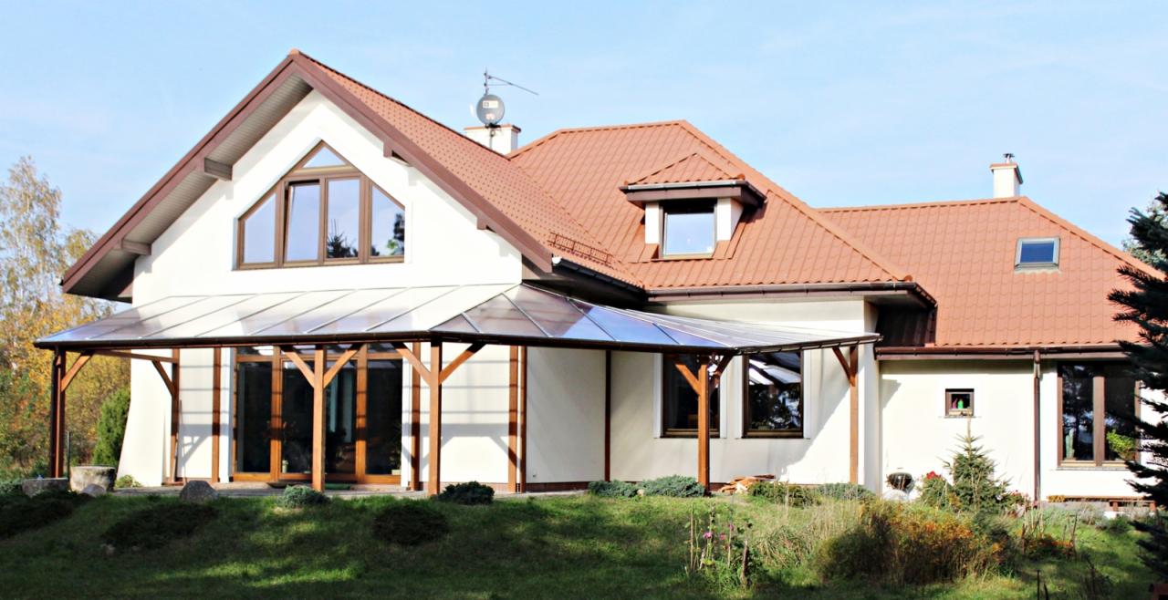 Zadaszony taras z poliwęglanu przy białym domu z brązowym dachem, widok z ogrodu w słoneczny dzień.