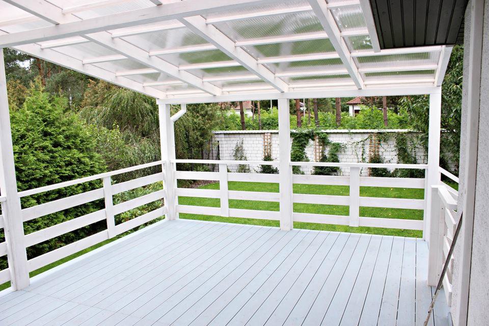 Zadaszony taras z poliwęglanu z białą drewnianą balustradą, szarymi deskami podłogowymi i widokiem na zielony ogród.