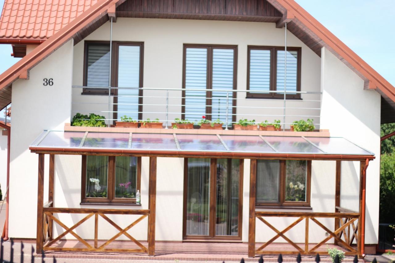 Zadaszony taras przy domu z balustradą balkonową, widoczne rośliny w donicach i drewniane elementy konstrukcyjne.