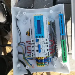 Otwarta biała skrzynka elektryczna z widocznymi przewodami, bezpiecznikami i sterownikiem na tle kostki brukowej.