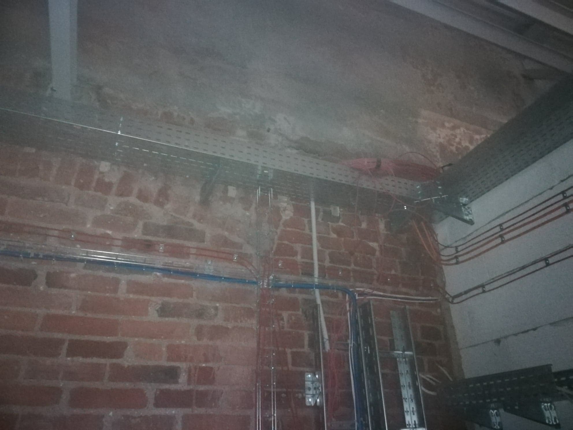 Surowe wnętrze z ceglaną ścianą, widoczne koryta kablowe i przewody elektryczne poprowadzone wzdłuż ściany i pod sufitem, w tle betonowy sufit.