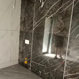 Wnętrze łazienki w trakcie remontu: ciemne płytki imitujące marmur na ścianie, jasne płytki na drugiej ścianie, szare płytki na podłodze, widoczne instalacje elektryczne i hydrauliczne, surowy stan...