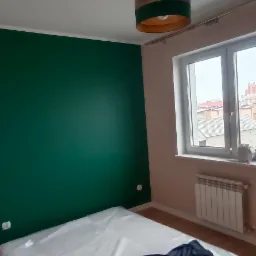 Pokój po odświeżeniu: ściana pomalowana na butelkową zieleń, zabezpieczone folią meble, okrągła lampa z brązowo-zielonym abażurem.