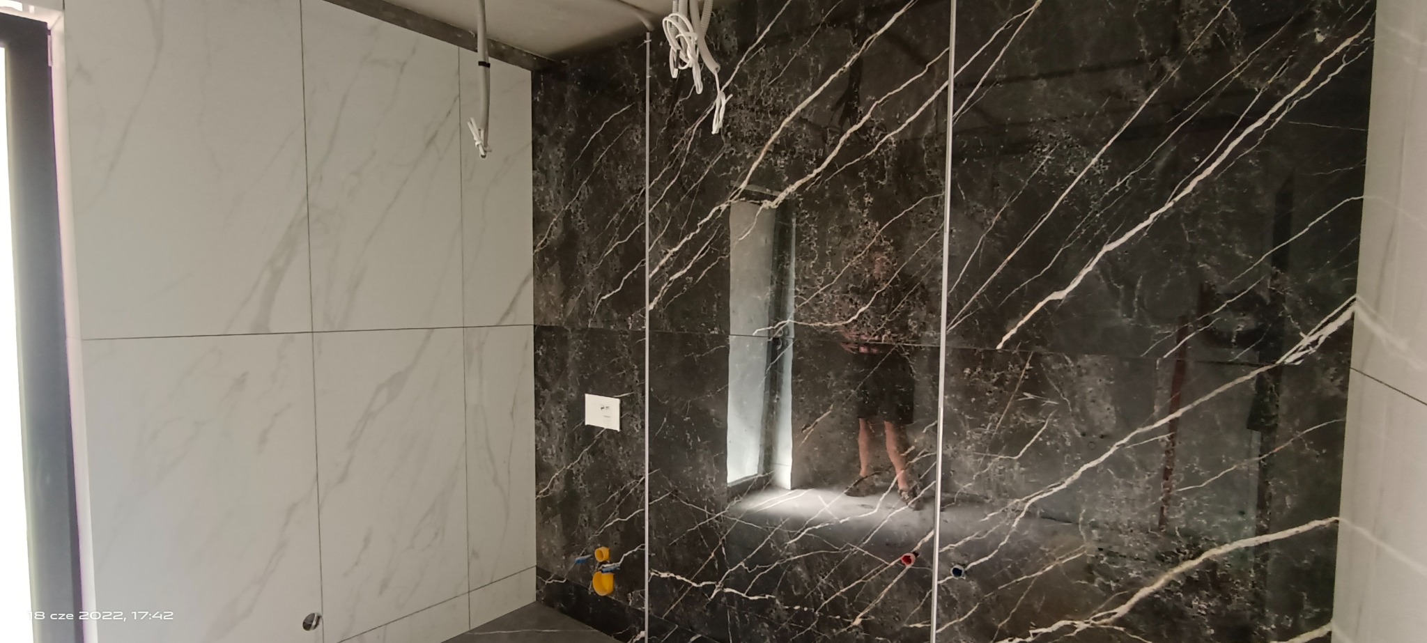 Częściowo wykończona łazienka z czarno-białymi płytkami imitującymi marmur, widoczne wystające kable i instalacja wodna, odbicie osoby w polerowanej powierzchni płytek.