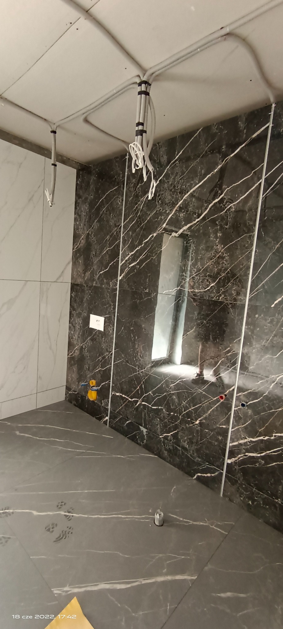 Wnętrze łazienki w trakcie remontu: ciemne płytki imitujące marmur na ścianie, jasne płytki na drugiej ścianie, szare płytki na podłodze, widoczne instalacje elektryczne i hydrauliczne, surowy stan...