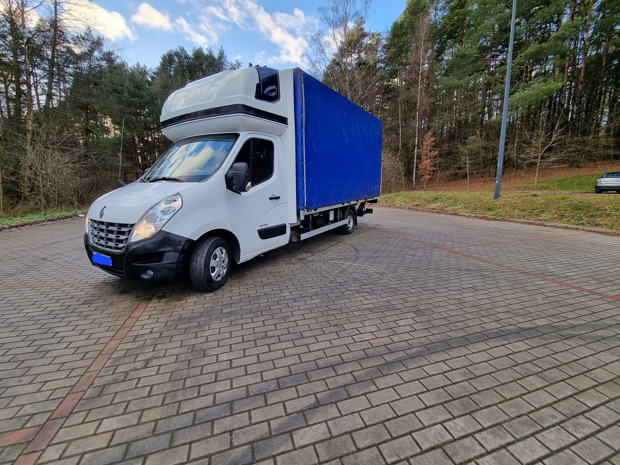 Biały samochód dostawczy Renault Master z niebieską plandeką na parkingu z kostki brukowej na tle lasu, widoczne drzewa bezlistne i iglaste, błękitne niebo z chmurami.