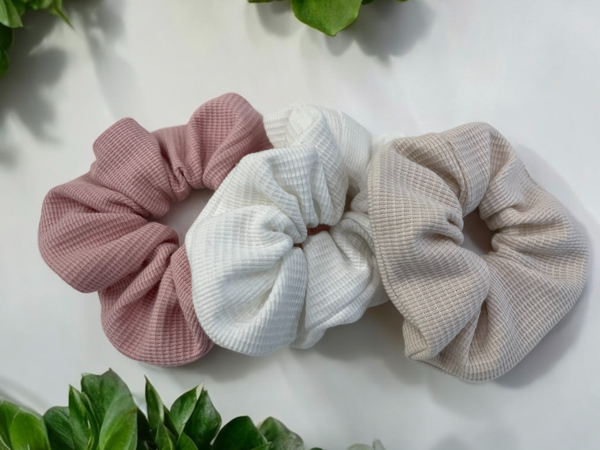 Trzy teksturowane gumki do włosów (różowa, biała, beżowa) leżą na białym tle, otoczone zielonymi liśćmi. Delikatne światło podkreśla fakturę materiału.